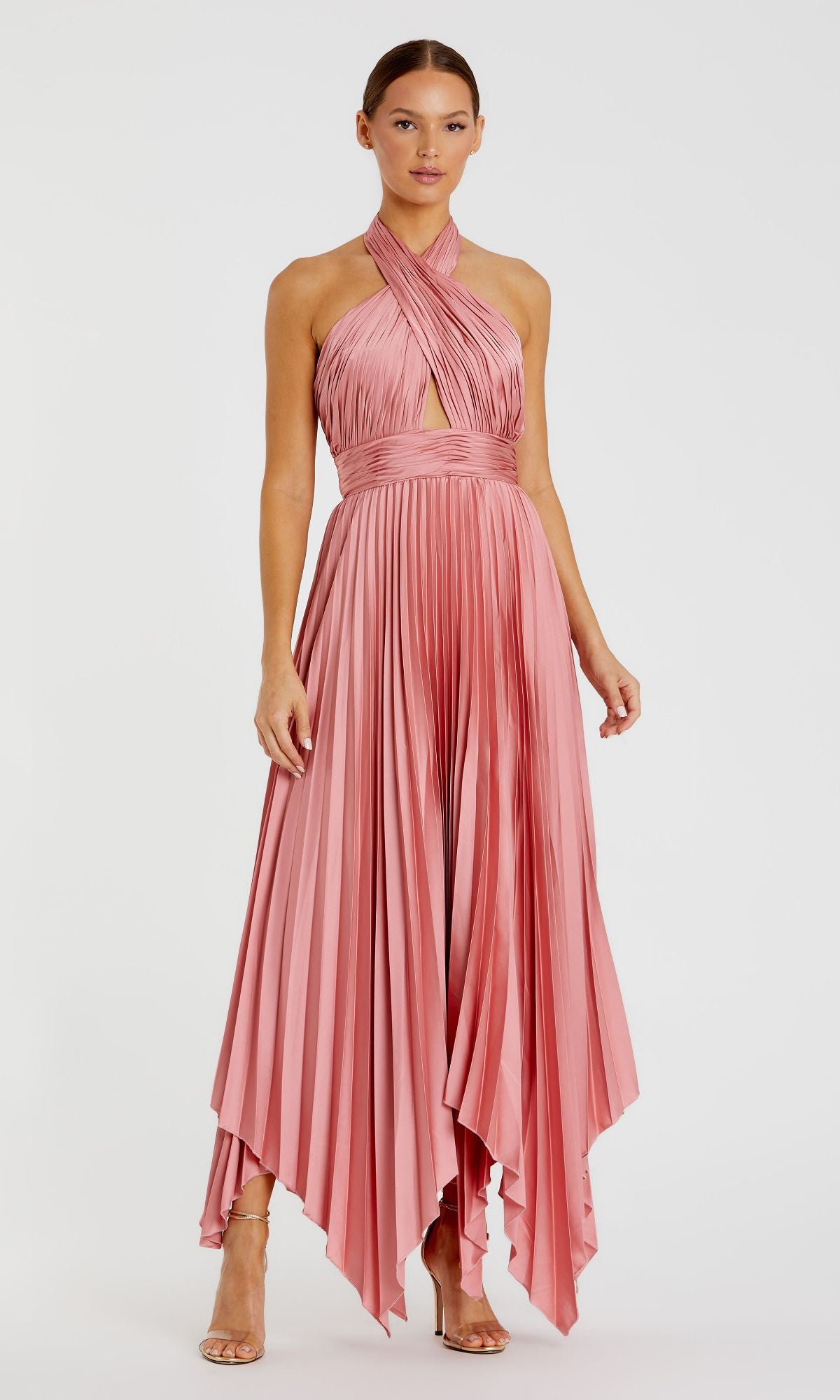 A woman stands against a plain white background wearing the Mac Duggal Long Formal Dress 11302— a sleeveless, pink, pleated halter gown with a handkerchief hem—paired with strappy heels.